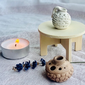 Può includere: Una piccola brocca in ceramica bianca con superficie testurizzata poggia su uno sgabello in legno chiaro. Accanto, un vaso in ceramica marrone con motivo a nido d'ape e aperture multiple riposa vicino a un lumino acceso e lavanda blu.