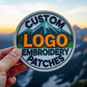 Puede incluir: Un parche bordado circular que presenta un gráfico de paisaje montañoso con árboles verdes y un pico azul. El texto CUSTOM LOGO EMBROIDERY PATCHES está cosido con hilo azul oscuro y naranja sobre un fondo blanco con un borde gris claro.