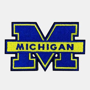 May include: A blue and yellow embroidered patch in the shape of a block letter M. A horizontal yellow bar runs across the center of the letter with the word MICHIGAN stitched in blue capital letters. The entire design has a yellow border.