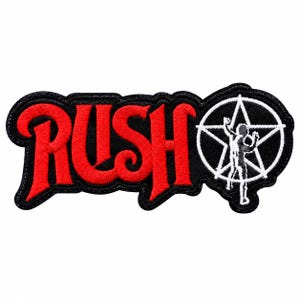 May include: An embroidered patch featuring the word RUSH in bold red lettering with a black border. To the right of the text is a white circular emblem containing a five-pointed star and a silhouette of a person standing with arms raised.