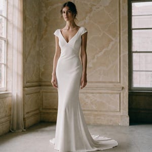 Peut inclure: Une robe longue blanche unie dotée d'un décolleté en V profond, de manches courtes et d'un corsage ajusté qui se prolonge par une jupe évasée avec une traîne. Le vêtement est confectionné dans un tissu mat et lisse aux lignes épurées.