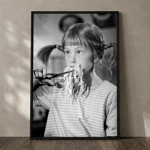 May include: A black and white framed wall art print featuring a young person with pigtails wearing a striped shirt. The person is holding a pair of scissors near their mouth, with a cluster of thin, noodle-like strands hanging from their lips.