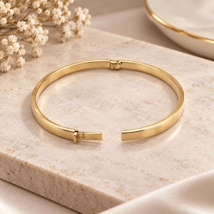Puede incluir: Una pulsera rígida de metal en tono dorado pulido con una superficie lisa y plana y un mecanismo de cierre con bisagra. La pulsera circular se muestra parcialmente abierta, revelando la bisagra integrada y el sistema de cierre.
