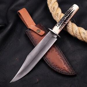 Puede incluir: Un cuchillo de hoja fija con una hoja de acero pulido y un mango de asta texturizado, que presenta una guarda de metal. El cuchillo se acompaña de una funda de cuero marrón con estampado decorativo en los bordes y cierre de broche.