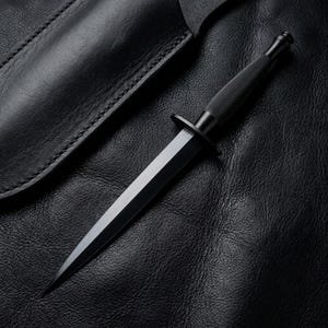 May include: A matte black double-edged dagger featuring a pointed blade, a circular crossguard, and a textured, knurled grip handle. The dagger is positioned alongside a matching black leather sheath with visible stitching along the edge.