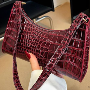May include: A deep burgundy shoulder bag with a glossy, embossed crocodile skin texture. The bag features a curved top silhouette, a single matching strap, and a zippered closure. A small circular logo with the text ZB ZOREVRABAGS is visible.