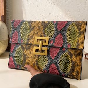 May include: A rectangular clutch bag featuring a vibrant snakeskin pattern in shades of yellow, pink, and grey. The bag is secured with a prominent, rectangular gold-toned metal clasp centred on the front flap, providing a structured closure.