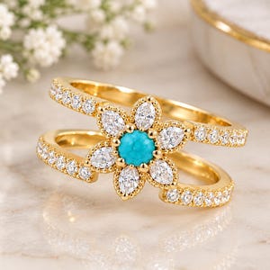 Peut inclure: Une bague ouverte dorée ornée d'un motif floral central avec une pierre turquoise ronde au milieu, entourée de six pierres claires taille marquise. Les deux anneaux fendus sont incrustés de petites pierres claires serties pavé.