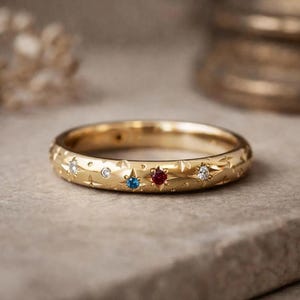 Peut inclure: Une bague en or avec une série de motifs en forme d'étoile gravés. La bague est ornée de petites pierres précieuses rondes et multicolores, dont une pierre bleue, une pierre rouge et plusieurs pierres blanches claires incrustées.