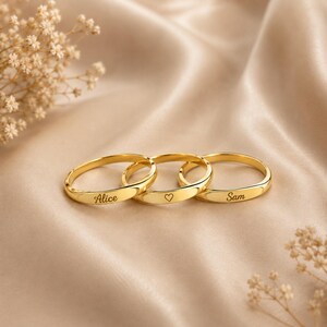 Peut inclure: Trois bagues en métal doré disposées en rangée. La première bague est gravée du prénom Alice, la deuxième présente un petit symbole de cœur et la troisième est gravée du prénom Sam. Chaque bague possède une finition lisse et polie.