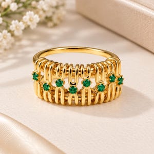 Puede incluir: Un anillo de oro con una banda ancha tipo jaula construida con bucles verticales entrelazados. El diseño está acentuado con pequeñas piedras preciosas redondas de color verde vibrante engastadas a intervalos a lo largo del centro.