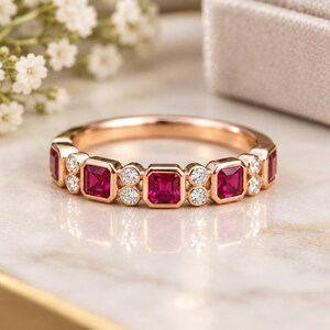 May include: A rose gold band ring featuring a repeating pattern of square-cut red rubies and pairs of small round white diamonds. The gemstones are set in a bezel style along the top half of the polished metal band, creating a symmetrical design.