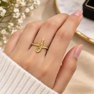 May include: A gold ring featuring a cursive letter J design with a single small clear gemstone set at the top terminal point of the letter. The band is smooth and polished, wrapping around the finger in a continuous, fluid, and open-ended shape.