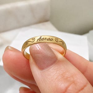 以下が含まれることがあります： 筆記体で「Aarau」という名前が刻印されたゴールドトーンのバンドリング。名前の両側には、装飾的な曲線を描く小さなハートのシンボルが刻まれており、パーソナライズされた細部までこだわったジュエリーアイテムとなっています。