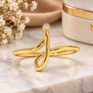 May include: A gold ring featuring a continuous, fluid, open-ended design that curves into a loop. A single, small, round, clear gemstone is bezel-set at the top terminal point of the polished gold band, creating a sleek and modern aesthetic.