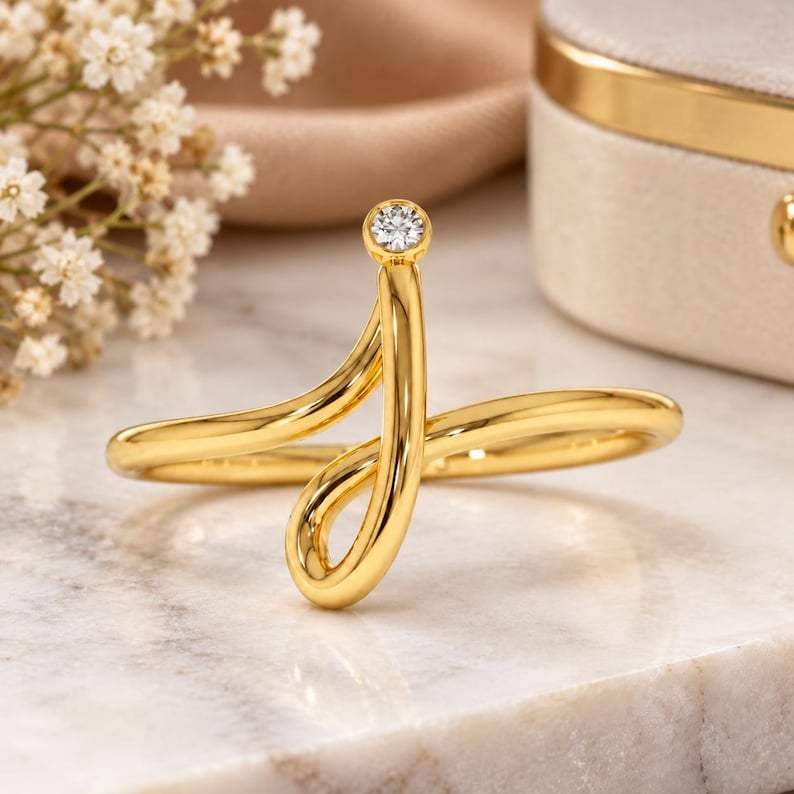 May include: A gold ring featuring a continuous, fluid, open-ended design that curves into a loop. A single, small, round, clear gemstone is bezel-set at the top terminal point of the polished gold band, creating a sleek and modern aesthetic.