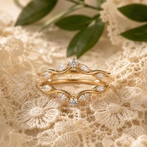 May include: A gold ring guard featuring two curved bands that meet at a central point. The bands are set with multiple marquise-cut and round-cut clear stones arranged in a symmetrical pattern along the polished gold metal frame.