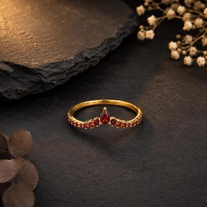 Peut inclure: Une bague en or dotée d'un anneau en forme de V, sertie de plusieurs petites pierres précieuses rouges rondes et d'une pierre rouge en forme de poire au centre. L'anneau présente une finition polie et une silhouette en chevron symétrique.