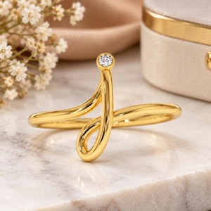 May include: A gold ring featuring a continuous, fluid, open-ended design that curves into a loop. A single, small, round, clear gemstone is bezel-set at the top terminal point of the polished gold band, creating a sleek and modern aesthetic.