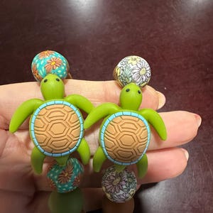 May include: Two beaded strands featuring green turtle-shaped charms with tan shells and light blue borders. The strands include round beads with floral patterns in teal, orange, white, and purple, alongside solid olive green and brown beads.