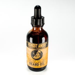 Op de afbeelding: Een bruine glazen fles met een zwarte druppelaar en een etiket met de tekst "Honest Amish" en "Pure Unscented Beard Oil". Het etiket bevat ook een cartoonafbeelding van een man met een baard en een hoed. De fles is 2 fl oz (59 ml).