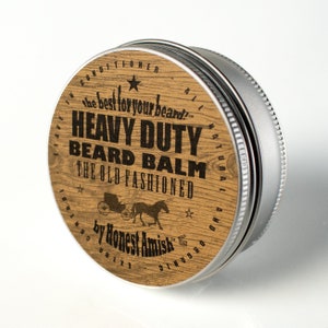Può includere: Un contenitore rotondo di metallo con un coperchio di legno. Il coperchio ha un motivo a venature di legno marrone e testo nero che recita "Heavy Duty Beard Balm The Old Fashioned by Honest Amish". C'è una piccola stella e un'immagine di una carrozza trainata da un cavallo sul coperchio.
