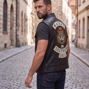 May include: A black leather vest featuring a large embroidered patch on the back with the text MAYANS, M.C., SOUTHERN, and CALI, along with a central graphic of a stylized skull mask. The vest is worn over a dark gray t-shirt and dark blue jeans.