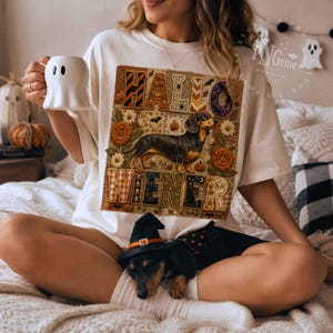 May include: An off-white t-shirt featuring a graphic of a dachshund surrounded by autumn flowers, pumpkins, and bats. The text reads "HALLOWIENER" in a blocky, patterned font. A small dachshund wearing a black witch hat sits in the foreground.