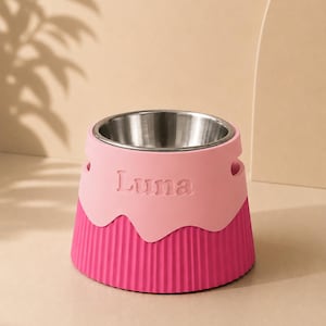 Pode incluir: Uma tigela para animais de estimação com um inserto de aço inoxidável e uma base em dois tons de rosa. A seção superior é rosa claro com o nome Luna em relevo na frente, enquanto a seção inferior é rosa magenta escuro com textura canelada.