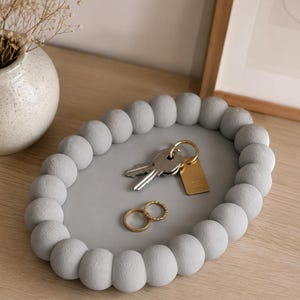 Pode incluir: Uma bandeja oval de cor cinza-claro com uma borda decorativa de esferas conectadas. A superfície central, lisa e plana, contém um conjunto de chaves de metal prateado num anel dourado com uma etiqueta retangular e dois anéis dourados.