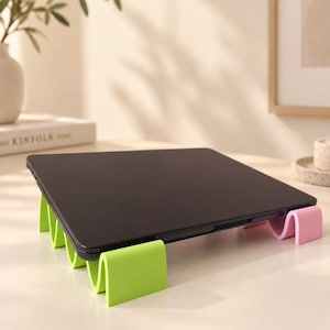 Puede incluir: Un ordenador portátil negro apoyado sobre dos soportes ondulados en forma de U. El soporte delantero es de color verde lima brillante, mientras que el trasero es de un suave rosa pastel. Los soportes presentan un patrón de ondas repetitivo.