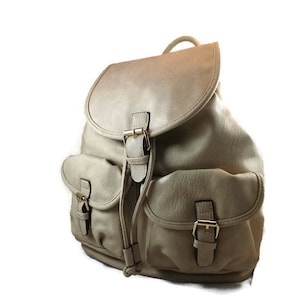 May include: A beige faux leather backpack with two front pockets and gold buckles. The backpack has a drawstring closure and adjustable straps.