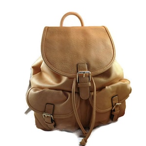 May include: A tan backpack with a curved flap and two front pockets. The bag has a top handle and adjustable straps. Buckles and drawstrings secure the bag's compartments. The backpack is made of a soft, textured material.