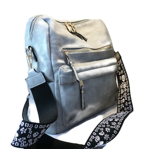 May include: A light gray faux leather backpack with a black and white patterned strap. The backpack has a zippered pocket on the front and a gold zipper pull.