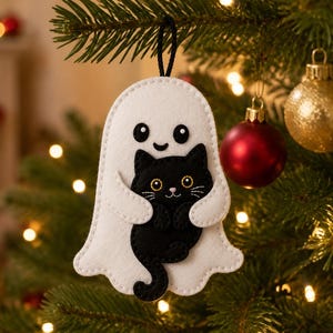 May include: A white felt ghost ornament featuring a smiling face and holding a small black cat with yellow eyes. The ghost has a black string loop at the top for hanging. The design is crafted with visible stitching along the edges of the felt.