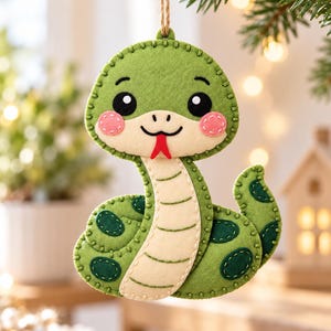 May include: A felt snake ornament in shades of green with a cream-colored underbelly, dark green oval spots, pink circular cheeks, black eyes, and a red forked tongue. The piece features visible blanket stitching along the edges and a twine loop.