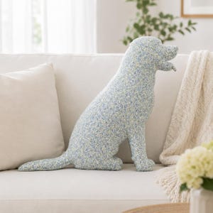 Puede incluir: Un cojín decorativo con forma de perro que presenta un estampado floral en azul claro y blanco. El cojín de tela relleno tiene la silueta de un perro sentado con cola, cuatro patas y la cabeza girada hacia un lado, con un diseño botánico denso.