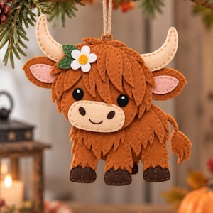 Op de afbeelding: Een vilten Highland-koe ornament in bruintinten met crèmekleurige hoorns, roze binnenoren en een witte bloem met groene blaadjes op de kop. De koe heeft donkerbruine hoeven, zwarte ogen en een gestikte glimlach, hangend aan een jute touwtje.