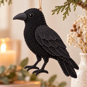 May include: A black felt crow ornament featuring visible grey blanket stitching along the edges, a small black bead for an eye, and a natural jute twine loop attached to the top for hanging. The bird is depicted in profile with layered wing details.