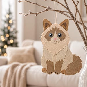 May include: A felt cat ornament featuring a cream and brown color palette with blue eyes, a pink nose, and stitched details. The cat is depicted in a seated position with a fluffy tail and is suspended by a thin string from a branch.