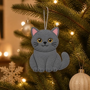 May include: A gray felt cat ornament featuring pink inner ears, large amber eyes, and a stitched nose and whiskers. The cat is in a seated position with its tail curled to the side and includes a silver hanging loop at the top of its head.