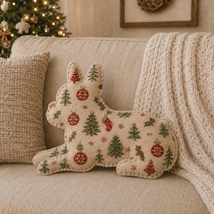 May include: A cream-colored throw pillow shaped like a French bulldog, featuring a repeating holiday pattern of green pine trees, red Christmas ornaments, small red stockings, and holly leaves with visible stitched edges along the silhouette.