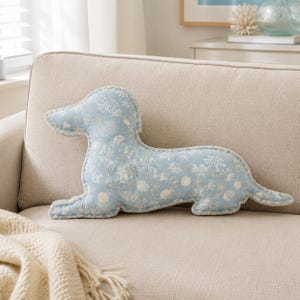 Pode incluir: Uma almofada em forma de cão salsicha na cor azul claro com um padrão repetitivo de conchas, estrelas-do-mar e motivos de coral em branco e bege. A almofada tem uma borda com debrum e uma estrutura macia e cheia, em forma de cão deitado.