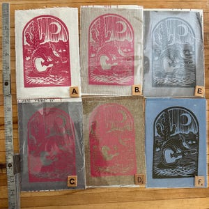 May include: A selection of fabric art prints depicting a desert scene with an armadillo playing a guitar, set beneath a crescent moon and cactus. The prints are in various colours, including red, grey, and blue, and are approximately 15 cm tall.