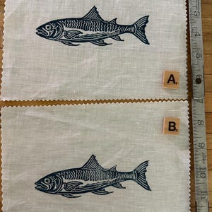 May include: Two off-white fabric squares featuring a dark blue fish print. The fish design is detailed, with scales and fins. The fabric has a raw edge. Wooden letter tiles labelled "A" and "B" are present.
