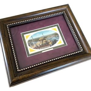 Op de afbeelding: Een donkerbruine houten fotolijst met een decoratieve kralenrand aan de binnenzijde, voorzien van een bordeauxrode passe-partout en een centrale rechthoekige print met een ovale afbeelding van een moskee met koepels en minaretten bij water.