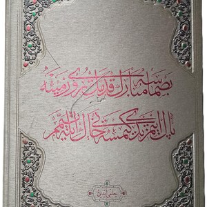 May include: A silver-colored hardcover book featuring intricate embossed floral corner patterns with small red and green accents. The center of the cover displays prominent magenta Arabic calligraphy, with a smaller rectangular cartouche containing additional text.