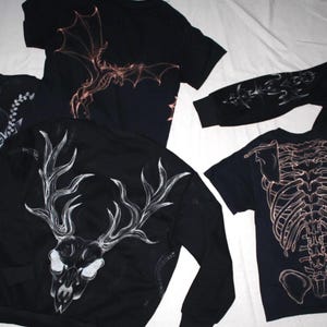 Könnte beinhalten: Eine Kollektion schwarzer Kleidungsstücke, darunter Kapuzenpullover und T-Shirts mit weißen und kupferfarbenen Grafikdrucken. Die Designs zeigen einen Hirschschädel mit Geweih, einen menschlichen Brustkorb, ein geflügeltes Wesen und florale Muster.
