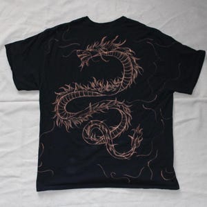 May include: A black short-sleeved t-shirt featuring a large, intricate, copper-coloured line art illustration of a serpentine dragon winding across the back. The design includes thin, flowing lines that extend towards the edges of the garment.