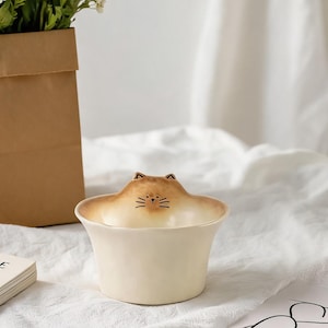 Könnte beinhalten: Eine cremefarbene Keramikschale mit einem geformten Katzenkopf an einer Seite. Die Katze hat braun gefärbte Ohrenspitzen, eine kleine Nase und schwarz aufgemalte Schnurrhaare. Die Schale hat einen glatten, abgerundeten Rand und einen konischen Boden.